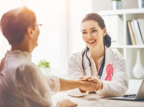 Doctor with pin ribbon on her lab coat sitting at her desk holding patients hand who is sitting across from her