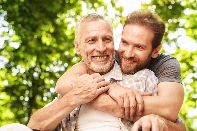 Son hugging father both are smiling there are trees in the background